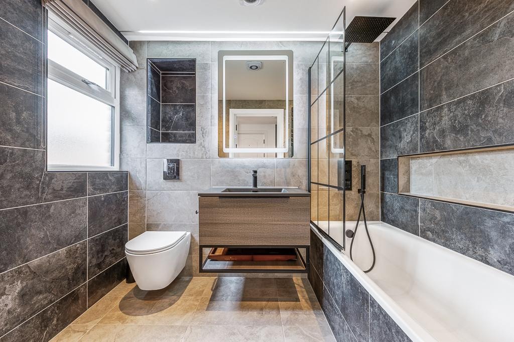 Melbourne Avenue — luxury bathroom with large-format dark marble-effect tiles, illuminated mirror, wall-hung WC and bath with shower
